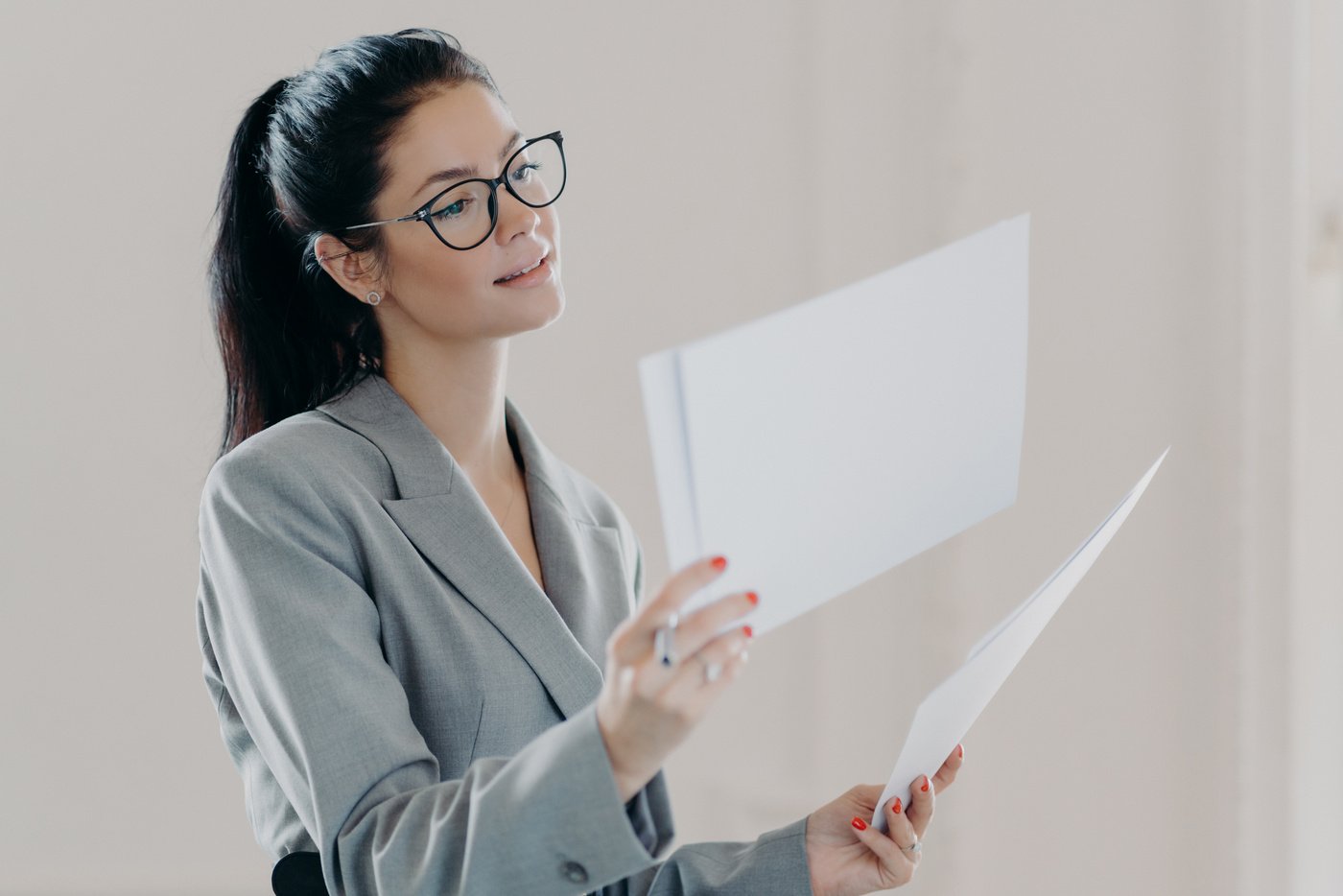 Photo of Female Financial Analyst Makes Report with Recommendations for Investments, Reads Strategy Information, Poses with Paper Documents, Dressed in Formal Clothes, Wears Eyeglasses, Stands Indoor